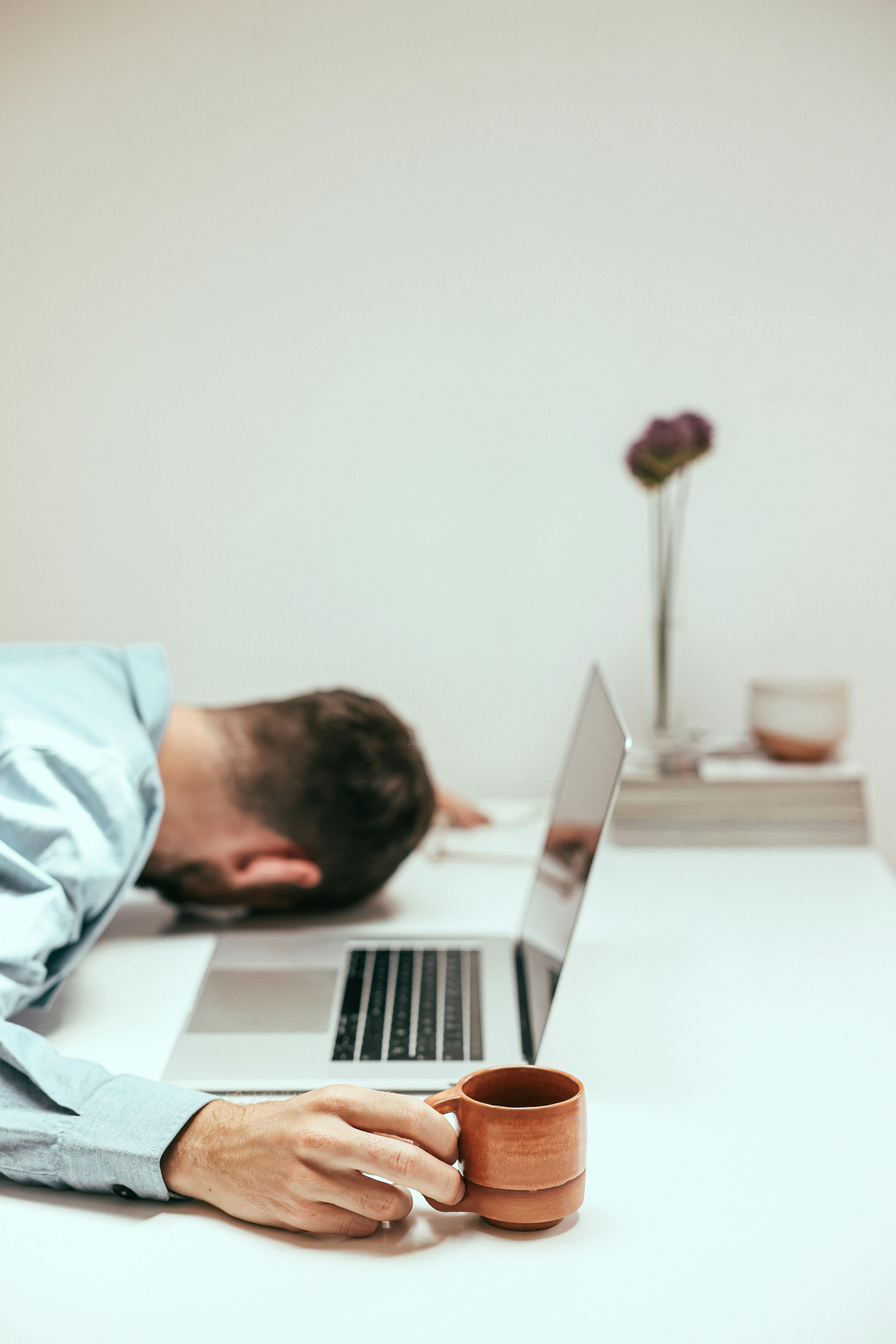 Stressed architect in New York with a coffee cup next to him, sleeping at his desk with his head on the table in front of his laptop.