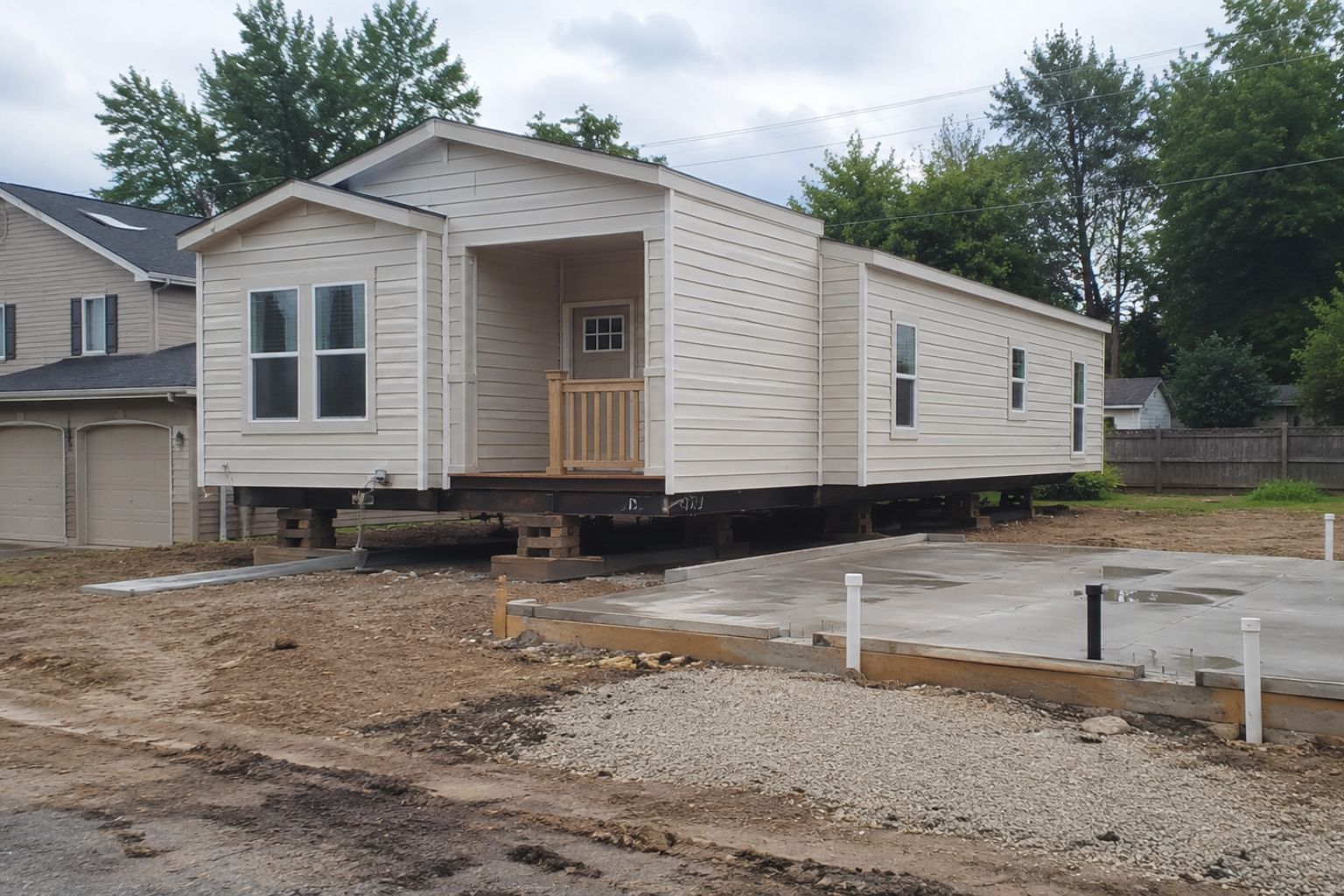 Exterior view of a single-wide manufactured mobile home being installed on a residential lot with concrete slab, utility pipes, and unfinished grading.