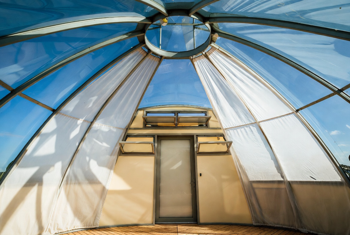 Glass dome house interior with curtains, overhead glazing, and visible openings for light and privacy control.