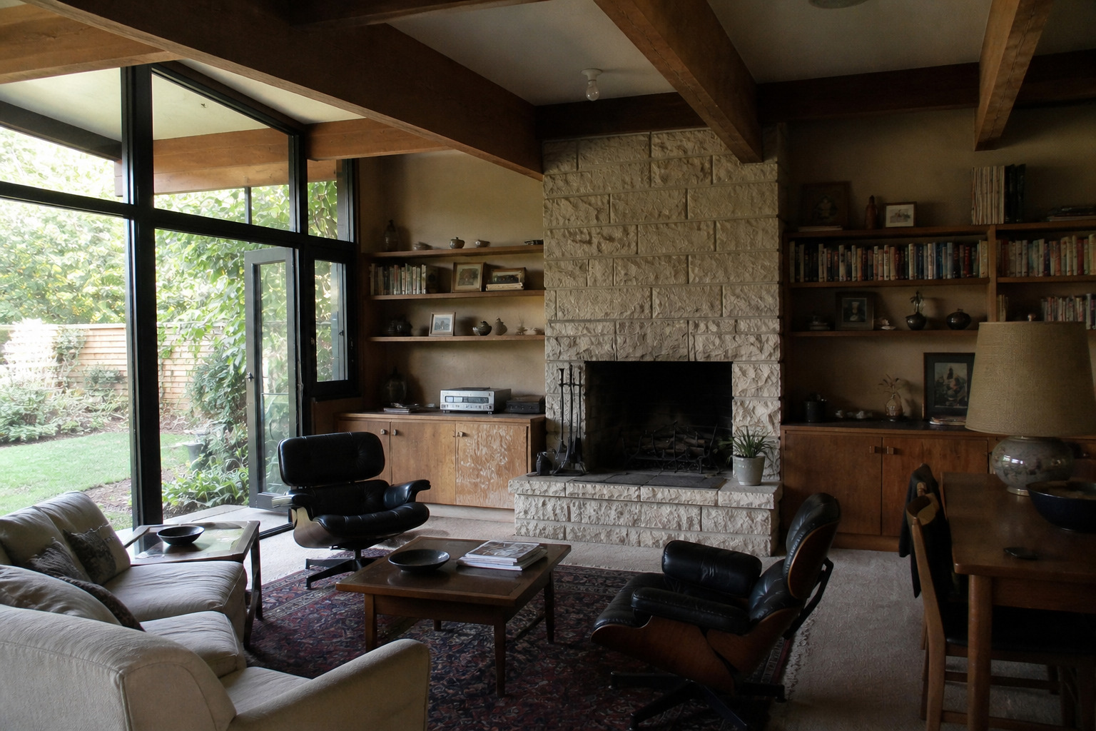 Mid-century living room with wood beams, stone fireplace, built-in shelves, large glass doors, and vintage lounge chairs.