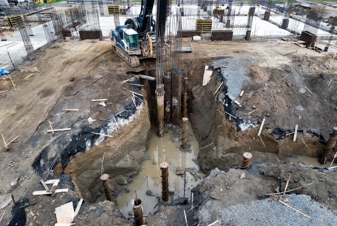 A crawler piling rig lowering a large rebar cage into a muddy drilled shaft, with helical piles installed nearby.