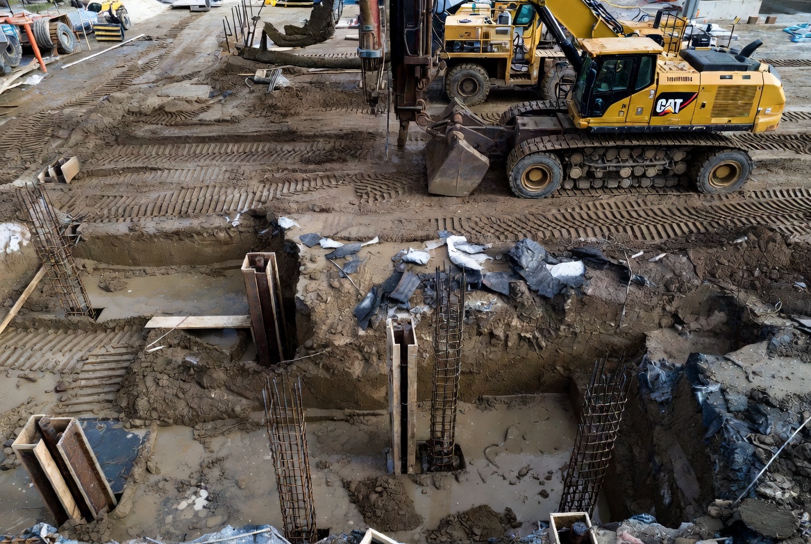 Deep foundation excavation with drilled piers, rebar cages, and muddy ground conditions.