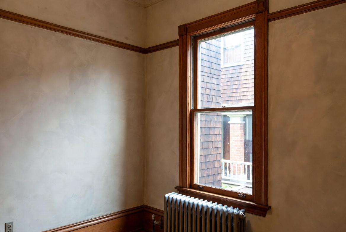 Interior corner of a 1910s house with plaster walls, wood trim, double-hung window, and cast-iron radiator.