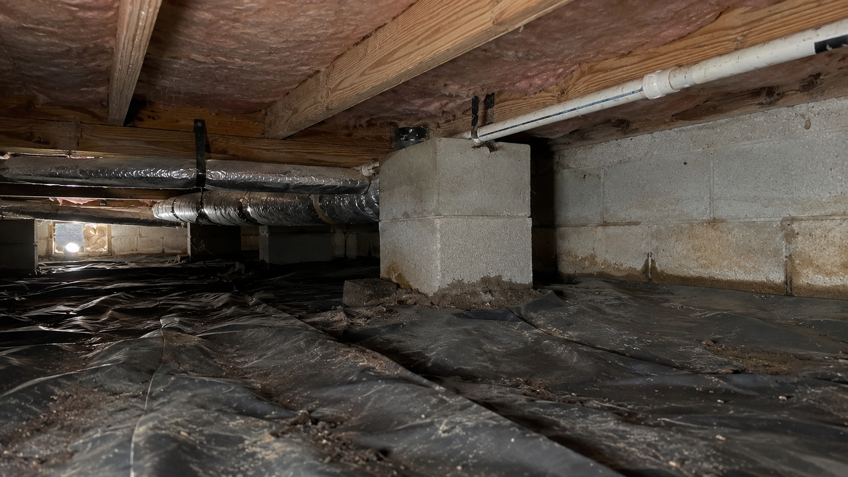 Low-clearance crawl space with ductwork, plumbing, block piers, and wrinkled ground liner that make inspection difficult.