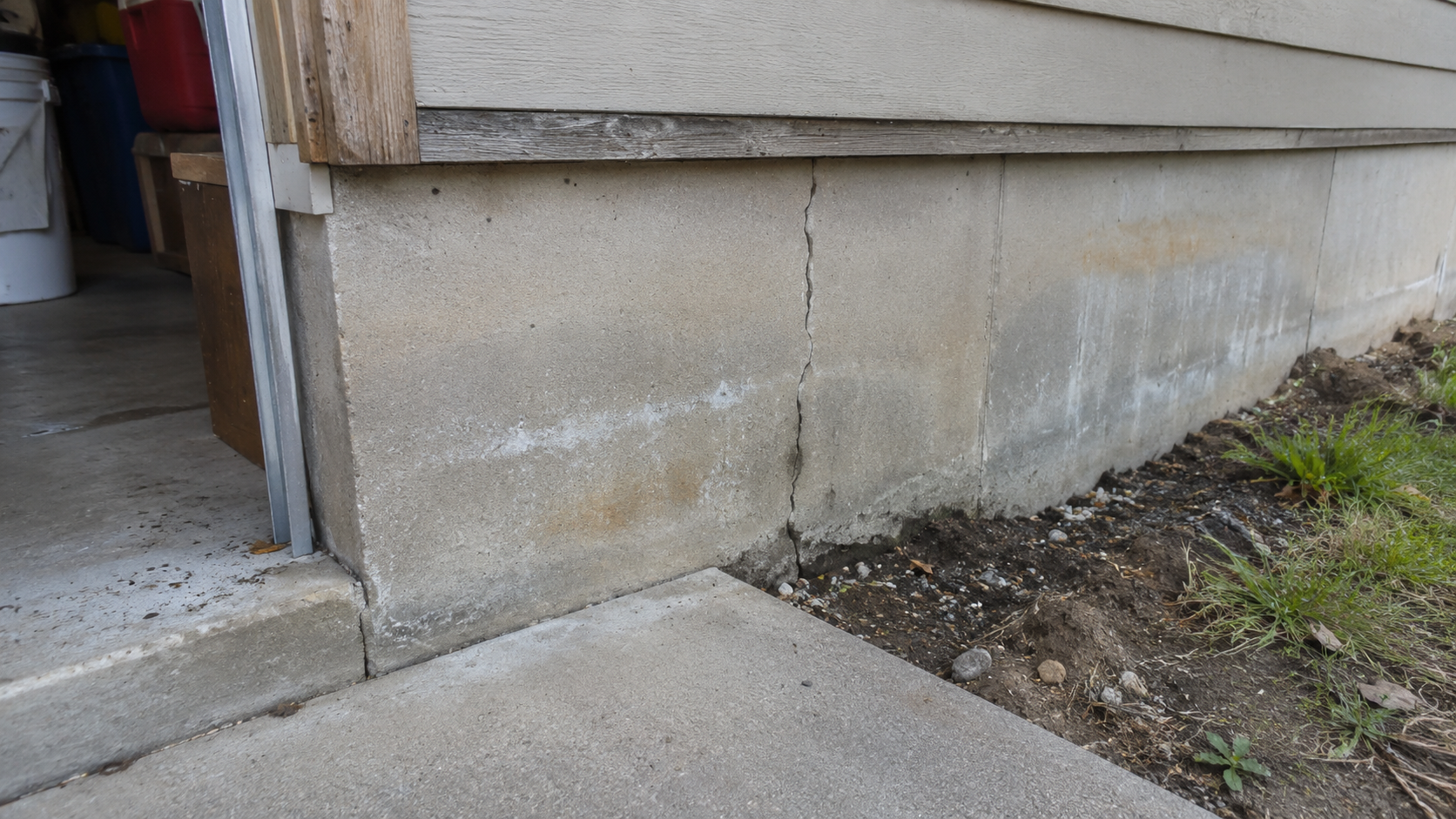 Cracked concrete stem wall beside a garage slab with water staining, soil exposure, and exterior grade visible along the foundation.