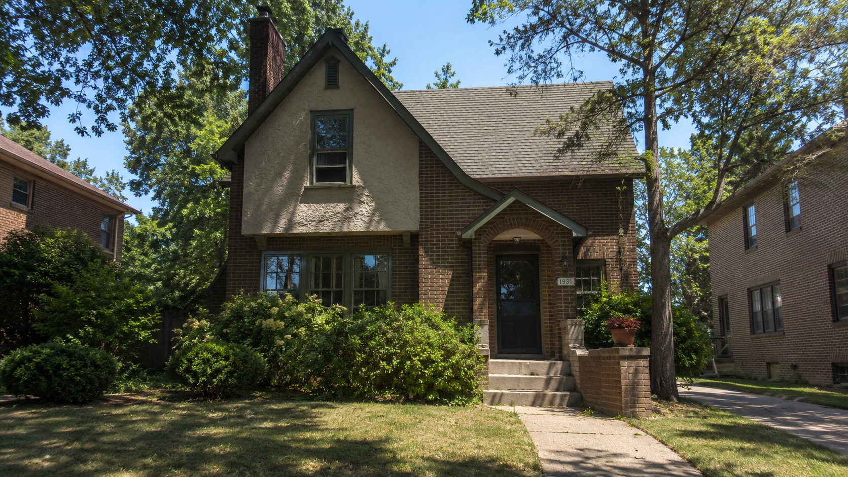 1930s brick and stucco house with steep gable, arched entry, mature shrubs, and front walkway.