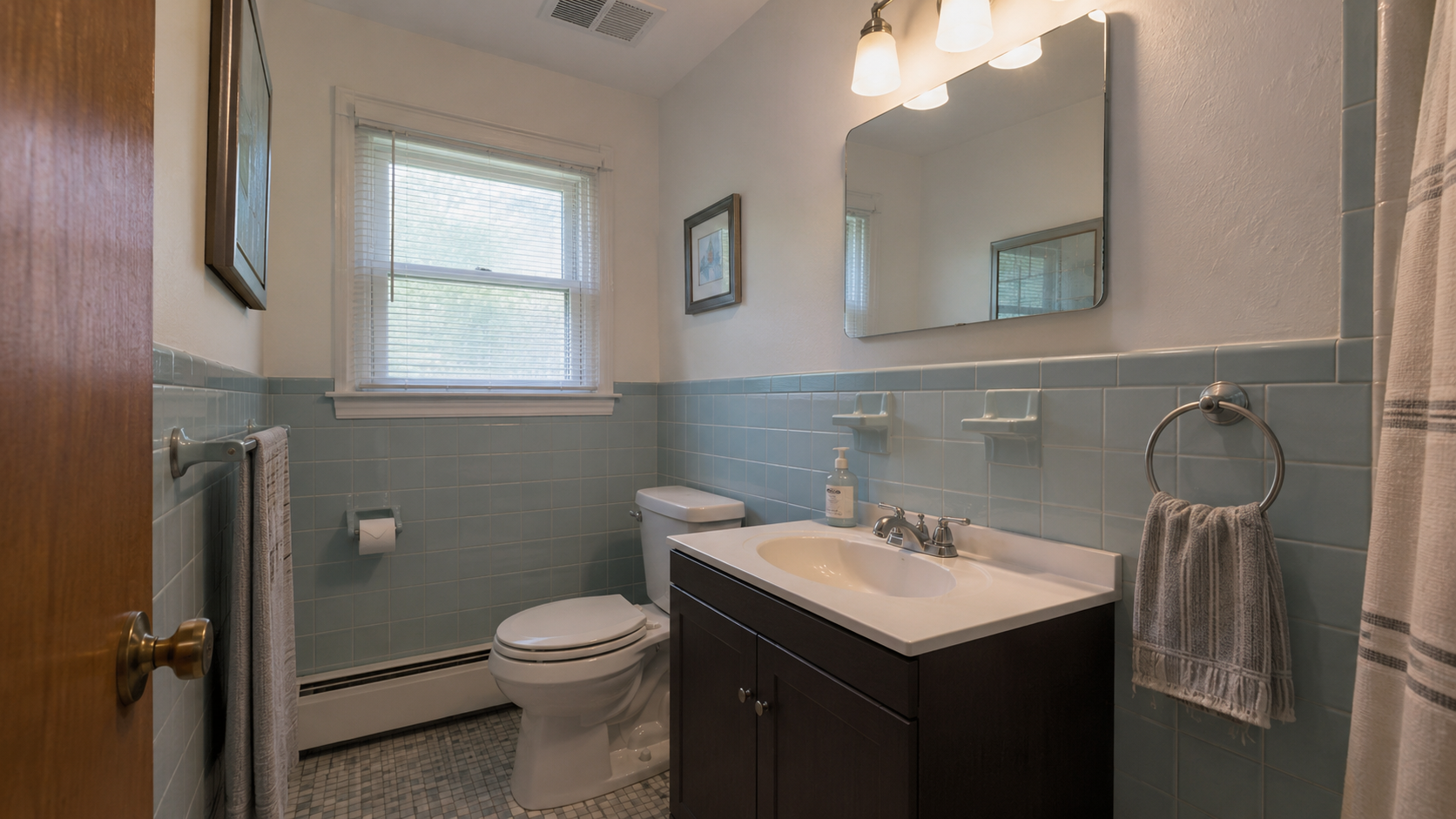 Modest 1960s bathroom remodel with original blue wall tile kept, updated vanity, mirror, lighting, fan grille, fresh paint, and repaired caulk.