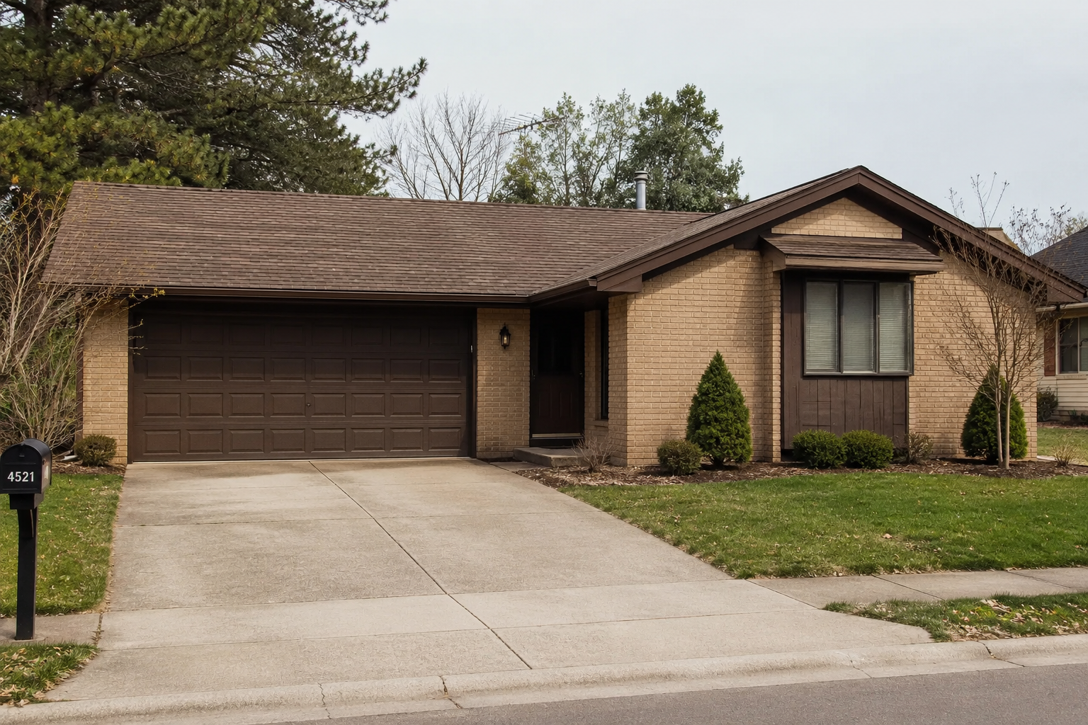 Modest 1980s suburban ranch house with beige brick, brown trim, front-facing two-car garage, recessed entry, driveway, and simple foundation planting.