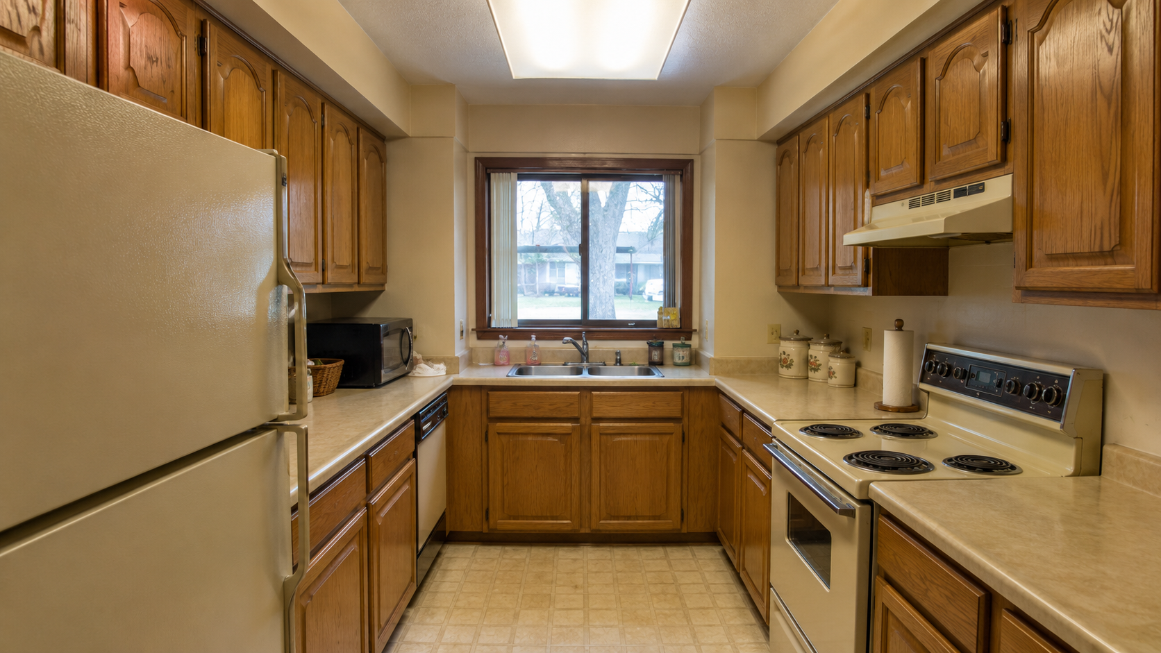 Untouched 1980s galley kitchen with oak cabinets, soffit, fluorescent ceiling light, and almond appliances.