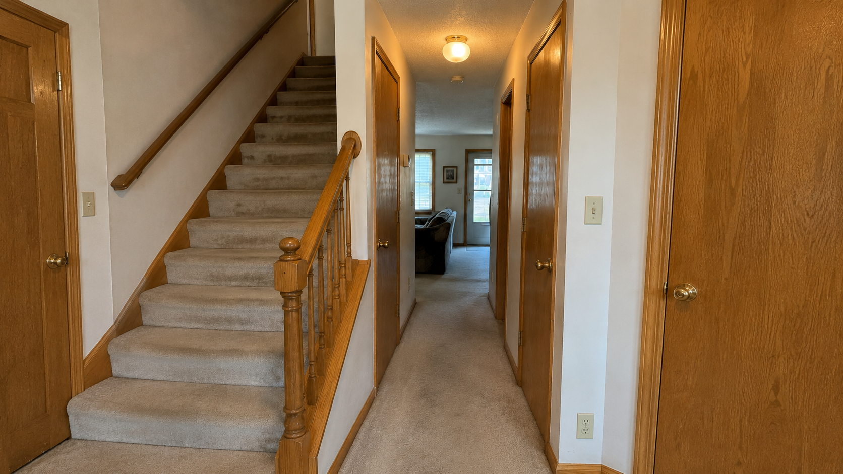 1980s hallway with oak stair rail, beige carpet, brass knobs, almond switches, hollow-core doors, and dated trim.