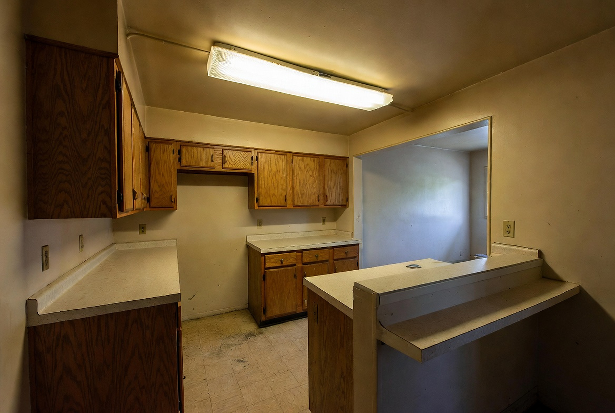 Original 1980s kitchen with oak cabinets, fluorescent light, peninsula counter, and low pass-through opening.