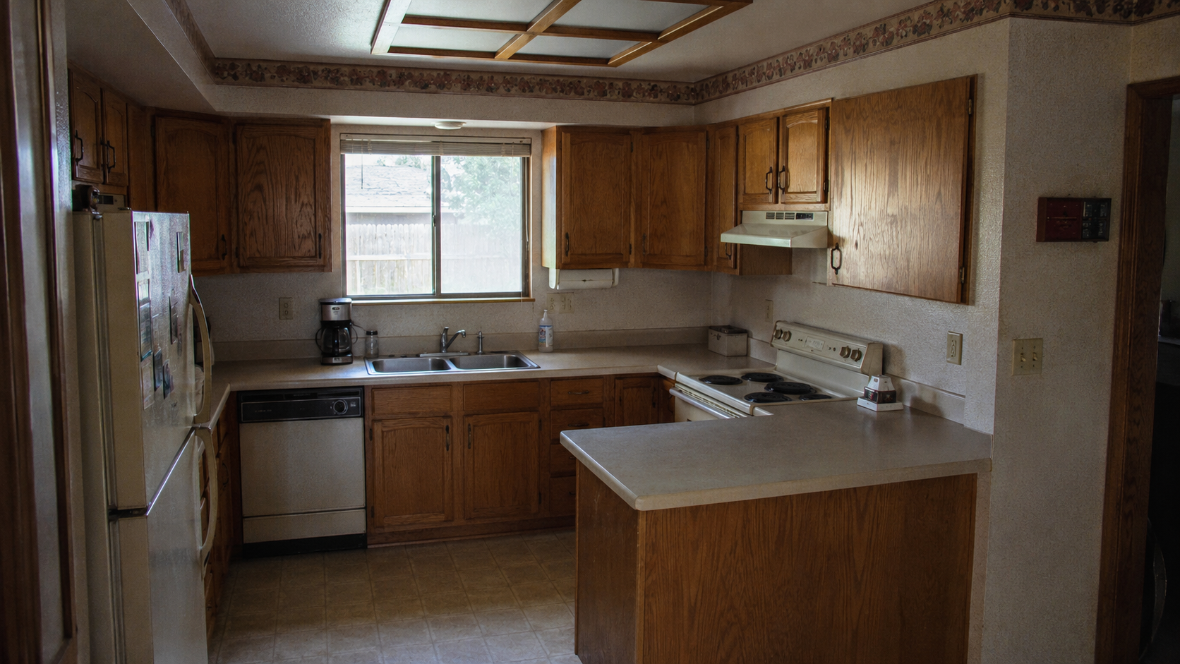 Modest 1980s suburban kitchen with honey oak cabinets, laminate counters, almond appliances, and fluorescent ceiling light.