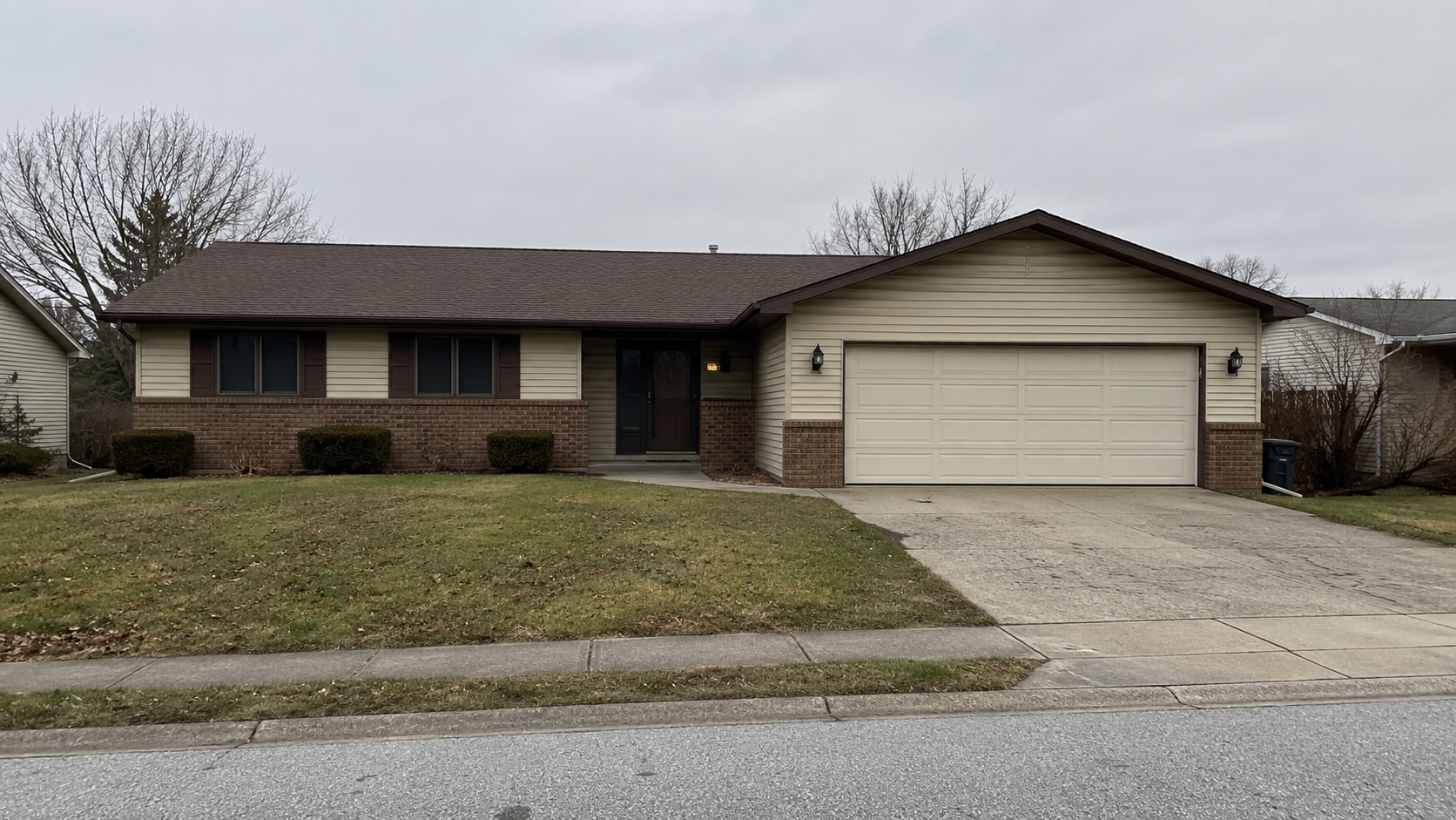 Modest 1983 suburban ranch house with beige siding, brown brick, front garage, patchy lawn, and low shrubs.