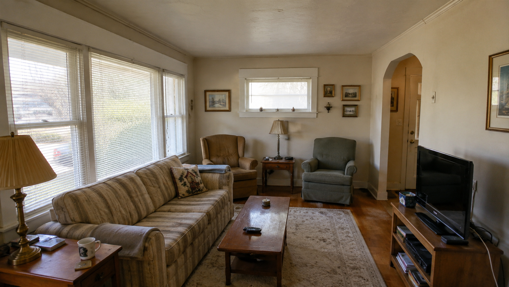 Original 1950s bungalow living room with modest furniture, large windows, and worn finishes.