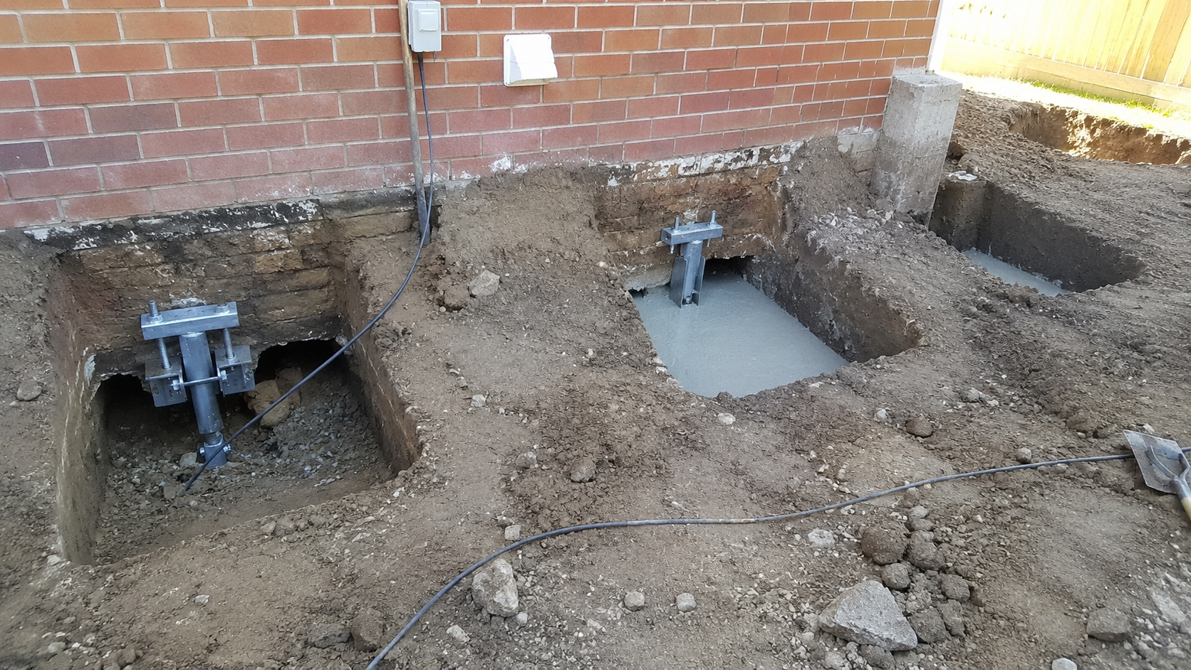 Residential foundation underpinning work in staged excavation bays beside a brick wall.