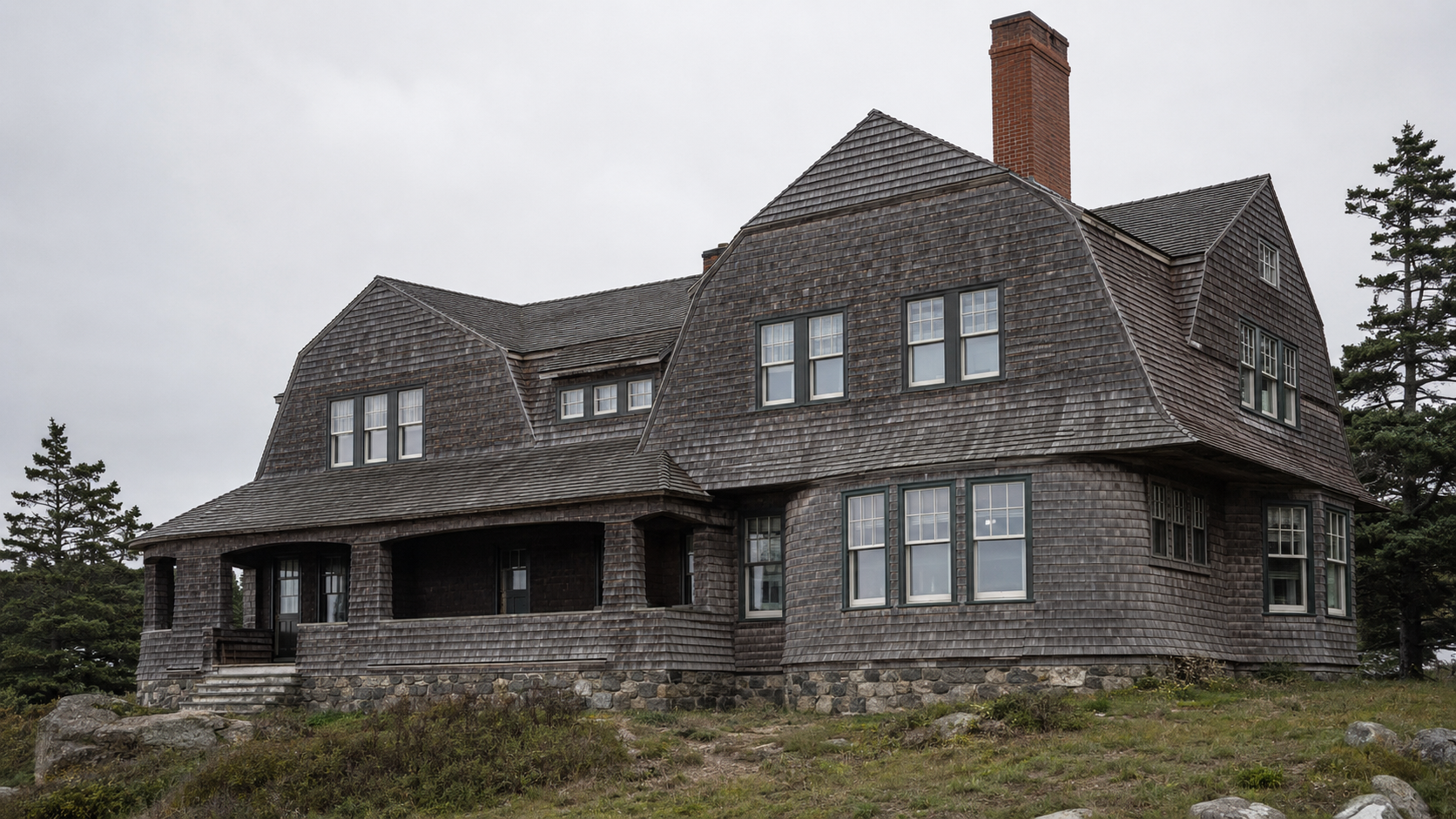 Weathered Shingle Style house with gambrel roofs, cedar cladding, porch, and stone base.