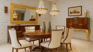 A dining room with stone accent wall, gold chandeliers, and cream upholstered chairs.