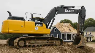 Excavator performing groundwork beside a house under active roof framing.