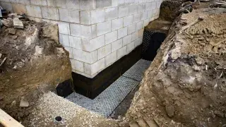 Exterior foundation waterproofing trench with drainage board, gravel, and coated wall.