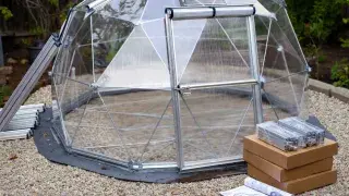 Greenhouse dome kit with visible frame, polycarbonate panels, door, and boxed components on a gravel base.