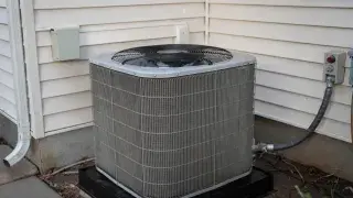 Outdoor residential heat pump condenser installed on a pad beside a house wall.