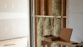 Careful mineral wool batt installation in open stud wall with cellulose and cork samples on workbench, premium construction detail.