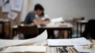 Architectural drawing tools, sketchbook, and plan sheets on a studio desk with students working in the background.