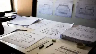 Architectural drawing sheets spread across a studio desk with plans and elevations pinned on the wall.