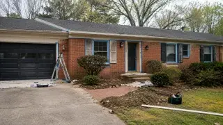 Brick ranch house exterior with taped trim, dark garage door, ladder, tools, and shrubs during an exterior update.