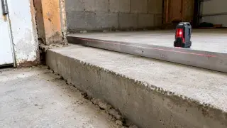 Low concrete slab edge with a visible crack and uneven elevation, shown with a straightedge and laser level in an unfinished garage or foundation area.