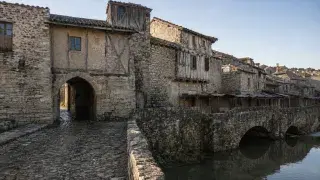 Medieval stone town edge with a gate passage, timber-framed upper floors, worn cobblestones, a riverside bridge, and repaired historic buildings.