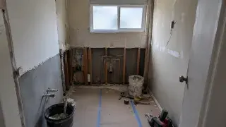Older ranch bathroom mid-remodel with exposed wall framing, rough plumbing, old tile, patched drywall, tools, and protected flooring.