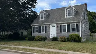 Documentary-style photo of a full Cape Cod house with cedar shakes, dormers, and a simple front facade.