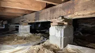 Pier and beam foundation repair inspection inside a crawl space with wood beams, stacked shims, masonry piers, dirt floor, and torn vapor barrier.