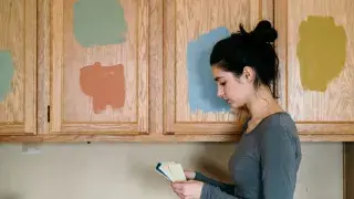 Woman comparing paint colors on oak kitchen cabinets with test swatches.
