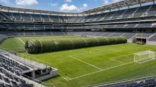 American-football field rolling away to reveal a natural-grass soccer pitch inside an existing stadium.