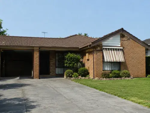 Close-up view of a modest 1980s brick suburban house with a low roofline, recessed entry, front driveway, awning, and foundation planting.