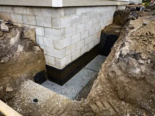 Exterior foundation waterproofing trench with drainage board, gravel, and coated wall.