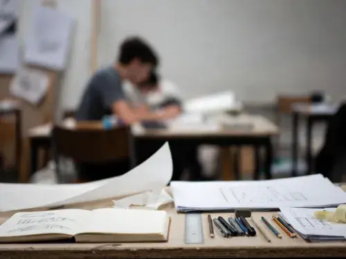 Architectural drawing tools, sketchbook, and plan sheets on a studio desk with students working in the background.