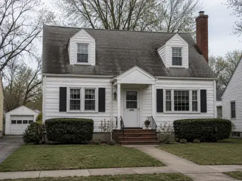 1940s Cape Cod-style house with white siding, dormers, brick chimney, and modest front entry.