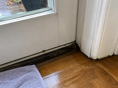 Wet interior floor at the bottom of an exterior door threshold, with water staining near the sill, worn trim, and a towel placed nearby after rain.