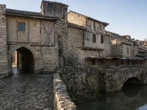 Medieval stone town edge with a gate passage, timber-framed upper floors, worn cobblestones, a riverside bridge, and repaired historic buildings.
