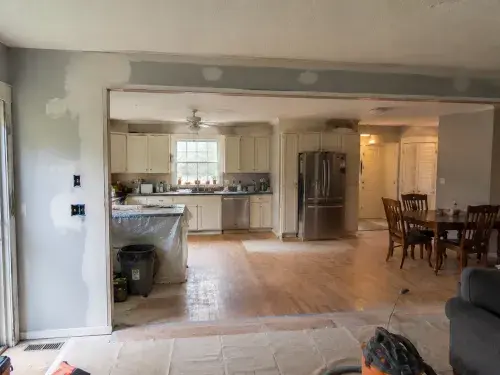 Older ranch kitchen opened to a dining and living area after wall removal, with patched drywall, floor changes, tools, and unfinished repairs.