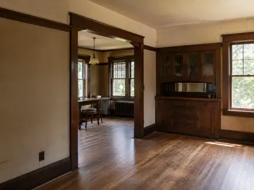 Original 1920s Craftsman interior with dark woodwork, built-in cabinet, radiator, and worn floors.