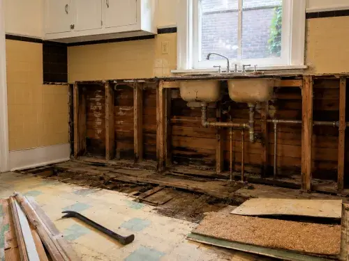 Open 1930s kitchen sink wall showing exposed plumbing, old wall boards, floor layers, and cabinet damage.