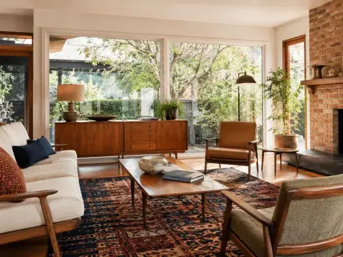Bright 1960s living room with warm wood furniture, patterned rug, brick fireplace, layered seating, plants, and bold accent pillows.