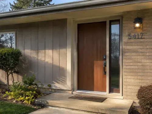 Updated 1970s front door with wood slab, sidelight, brick, modern hardware, and entry lighting. Caption A good 1970s front door tells you more than the door style; the glass,