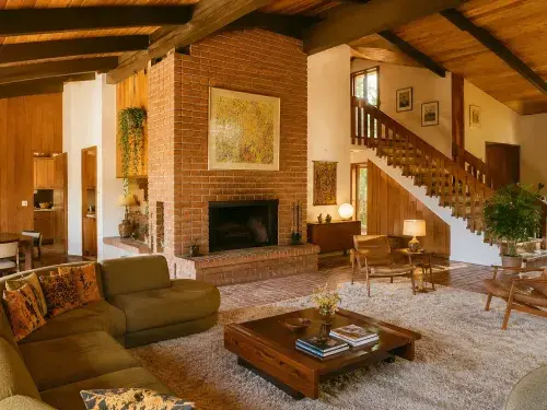 Bright 1970s living room with brick fireplace, wood ceiling, open stair, and low seating.