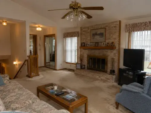 Dated 1980s family room with beige carpet, oak trim, brick fireplace, brass fan, vertical blinds, and mirrored doors.