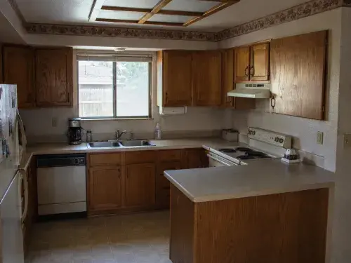Modest 1980s suburban kitchen with honey oak cabinets, laminate counters, almond appliances, and fluorescent ceiling light.